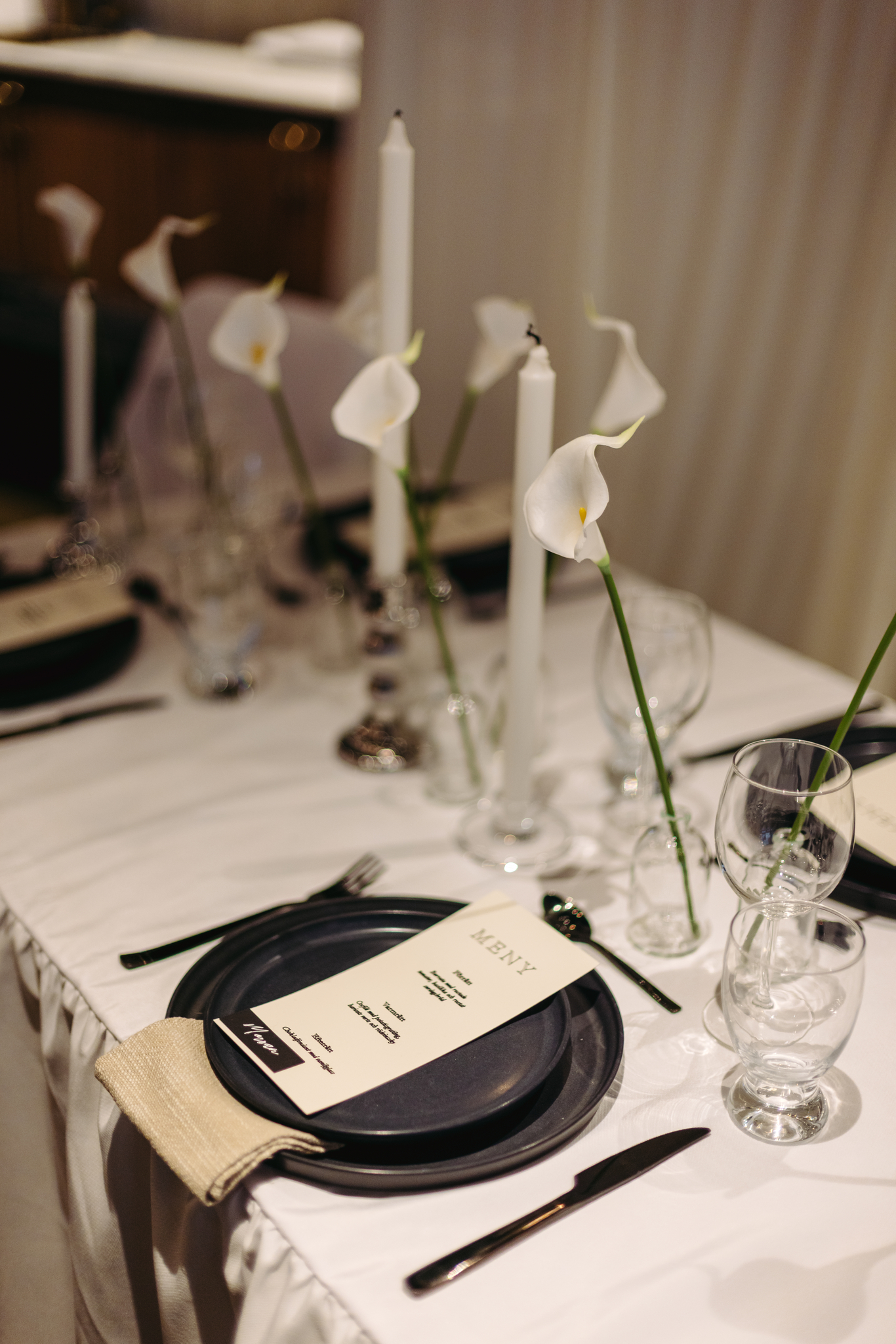 Elegant event table setting with black plates, tall white candles, and calla lilies by Timeless Moments AB